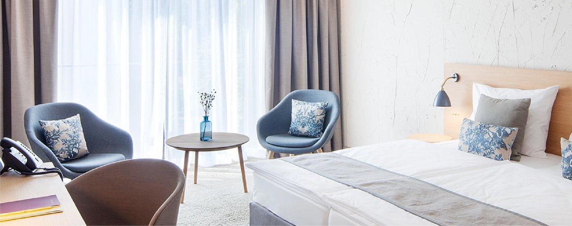 Camera d’hotel in toni neutri con tende bianche, poltroncine azzurre e cuscini coordinati