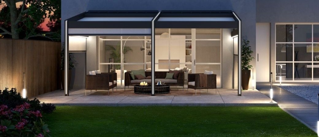 Pergola con vetrate scorrevoli davanti a una casa, illuminata di sera sul prato