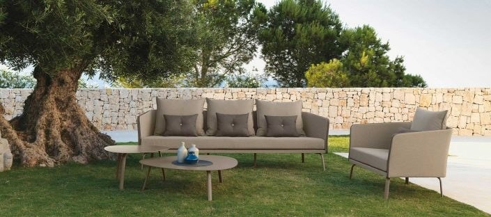Divano e poltrona beige con tavolini tondi sul prato, sotto un ulivo