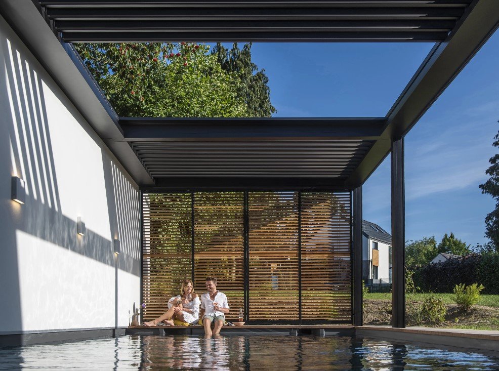 Pergola Skye a bordo piscina, con parete in legno a lamelle e una coppia seduta