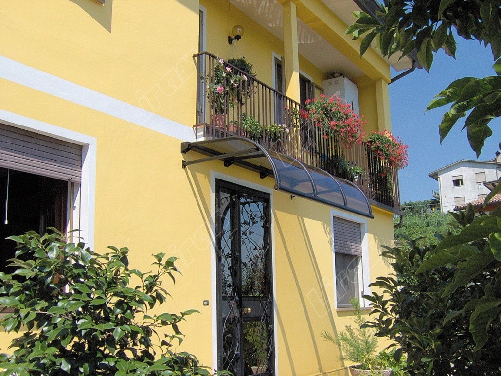 Pensilina a vela sotto un balcone fiorito, sopra una porta d’ingresso