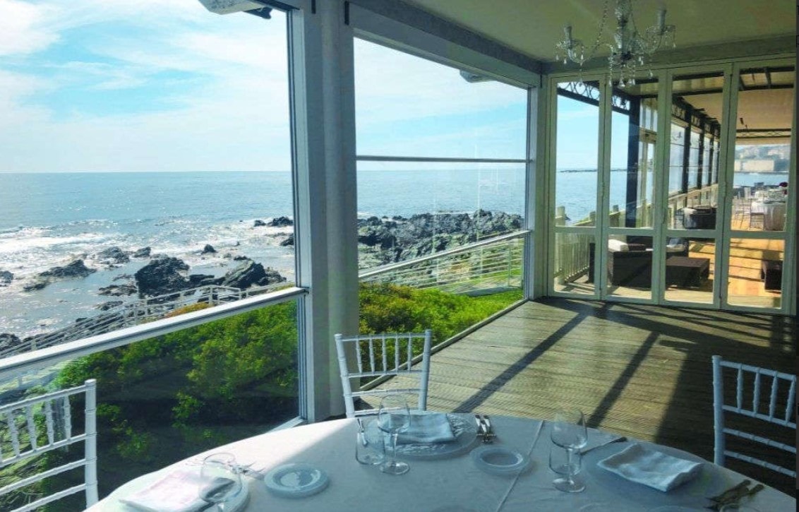 Terrazza di un ristorante sulla scogliera chiusa da vetrate saliscendi, con un tavolo apparecchiato