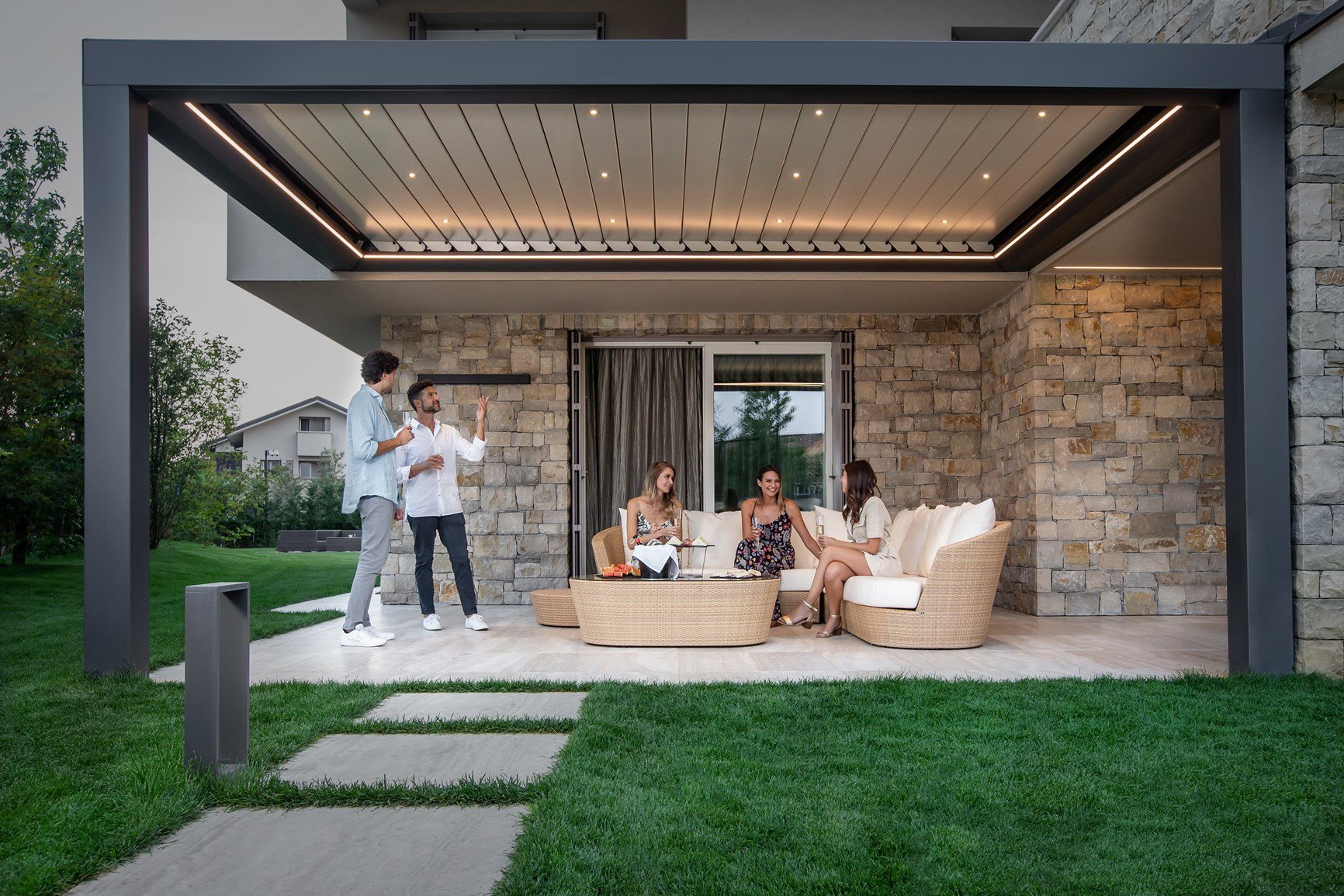 Pergola bioclimatica con luci integrate davanti a una casa in pietra, con una famiglia sul divano