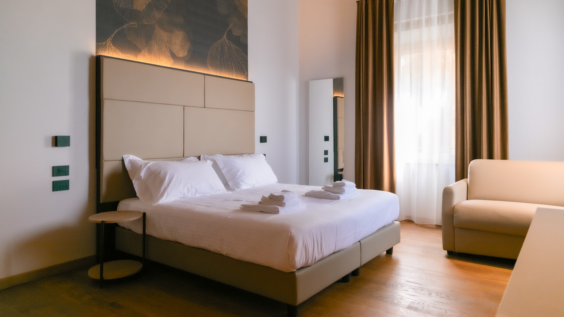 Camera d’hotel con letto matrimoniale, testiera imbottita retroilluminata e tende color bronzo