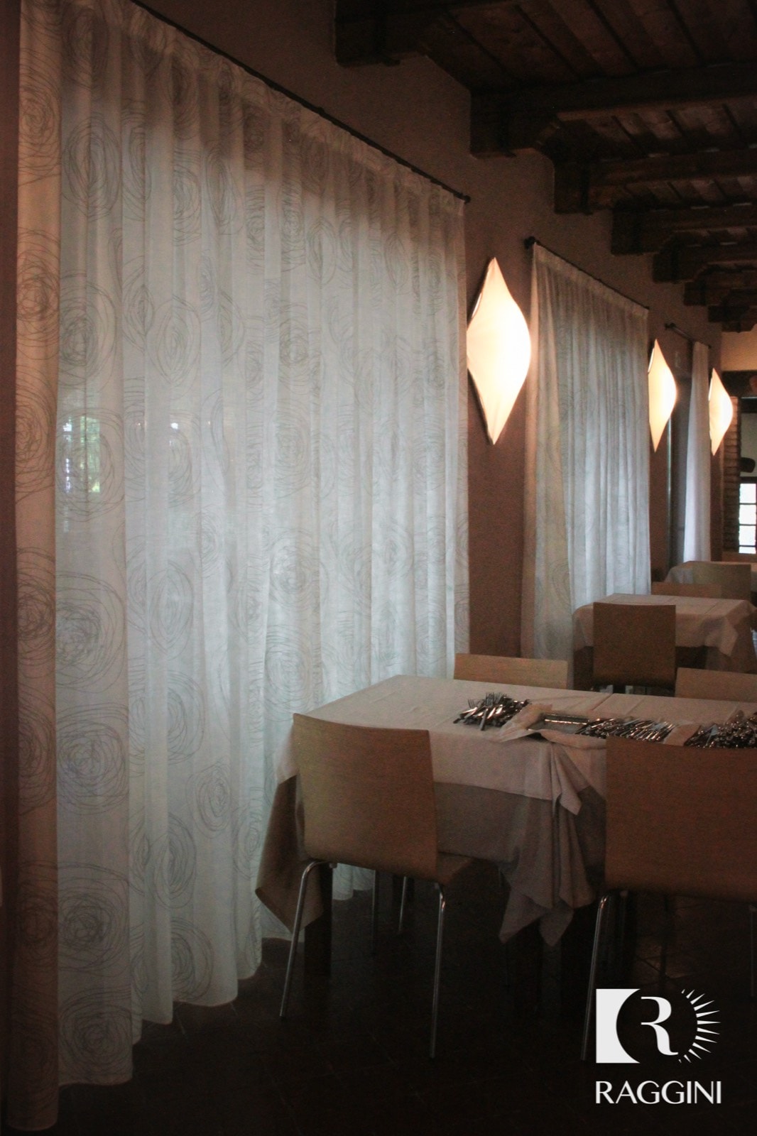 Tavoli apparecchiati e sedie imbottite nella sala di un ristorante, davanti a tende chiare