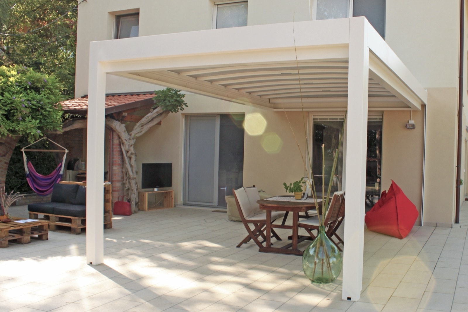 Pergola Maestro bianca con telo impacchettabile sul patio di una casa, con tavolo e pouf rosso