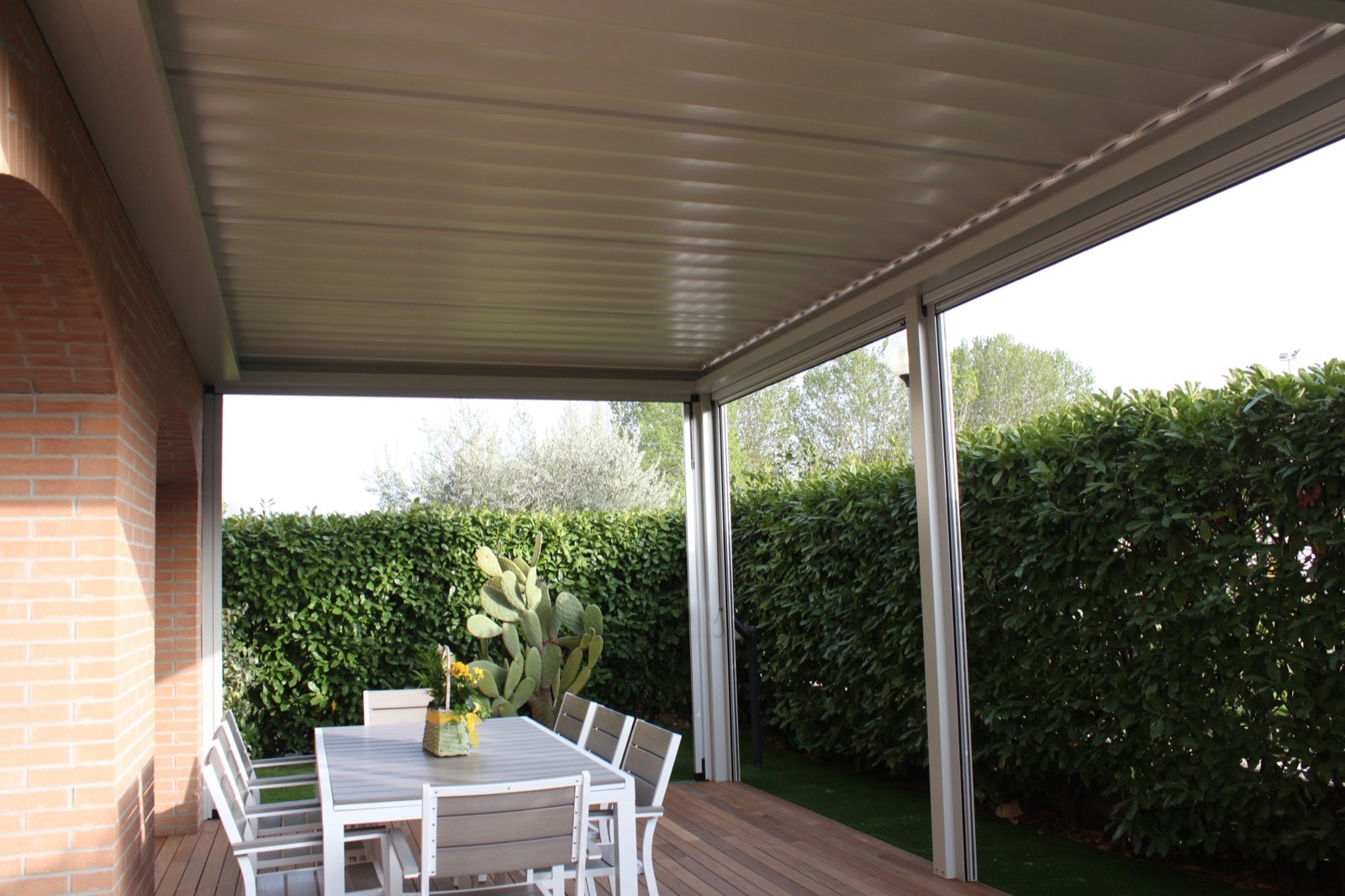 Pergola a lamelle grigia con tavolo da pranzo sulla pedana in legno, davanti a una siepe