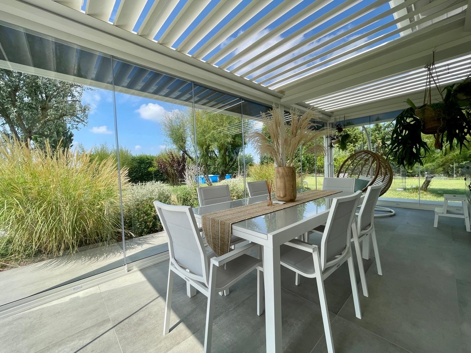 Pergola bioclimatica bianca a lamelle con tavolo da pranzo, affacciata su un giardino con graminacee