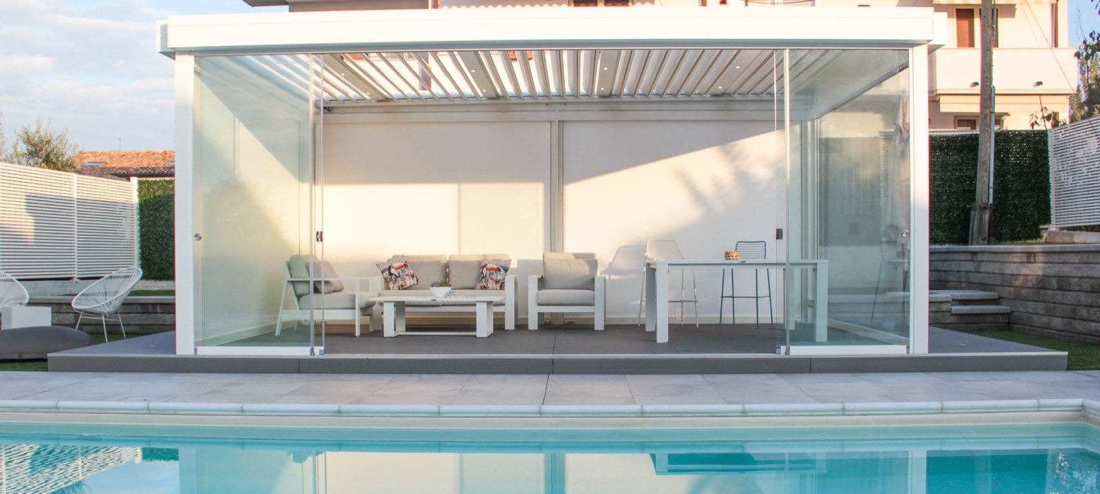 Pergola bioclimatica bianca con chiusure laterali in vetro accanto a una piscina