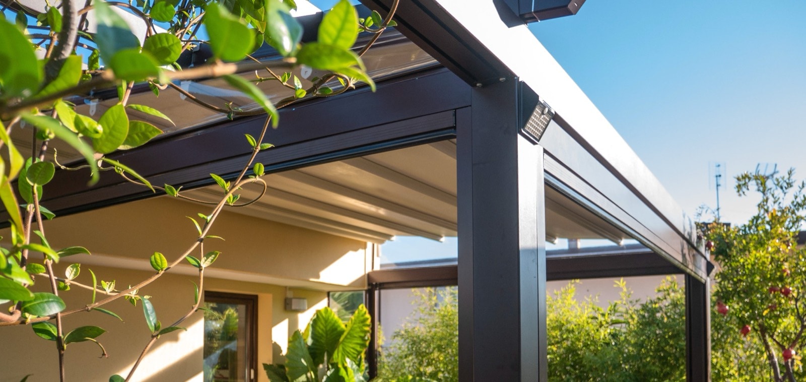 Pergola addossata in alluminio marrone con telo a impacchettamento, circondata da piante rampicanti