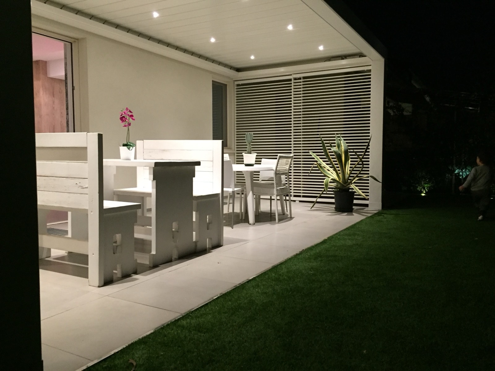 Pergola addossata bianca illuminata di sera con zona pranzo sul prato
