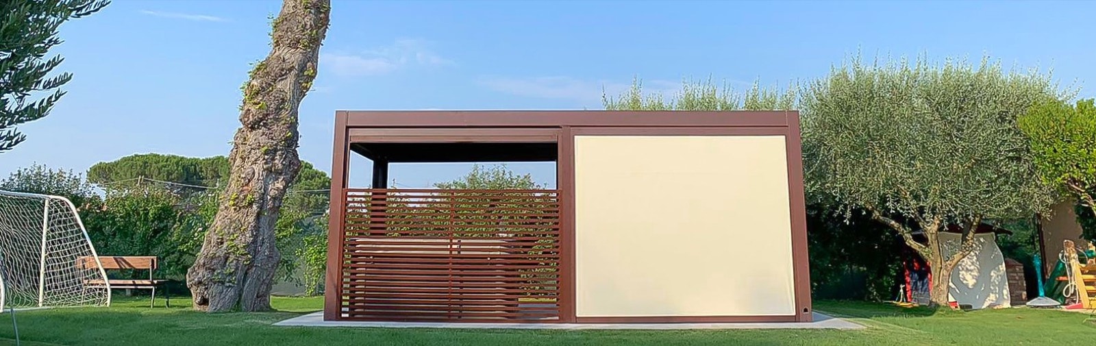 Pergola autoportante marrone con chiusure laterali in giardino, fra un pino e gli ulivi
