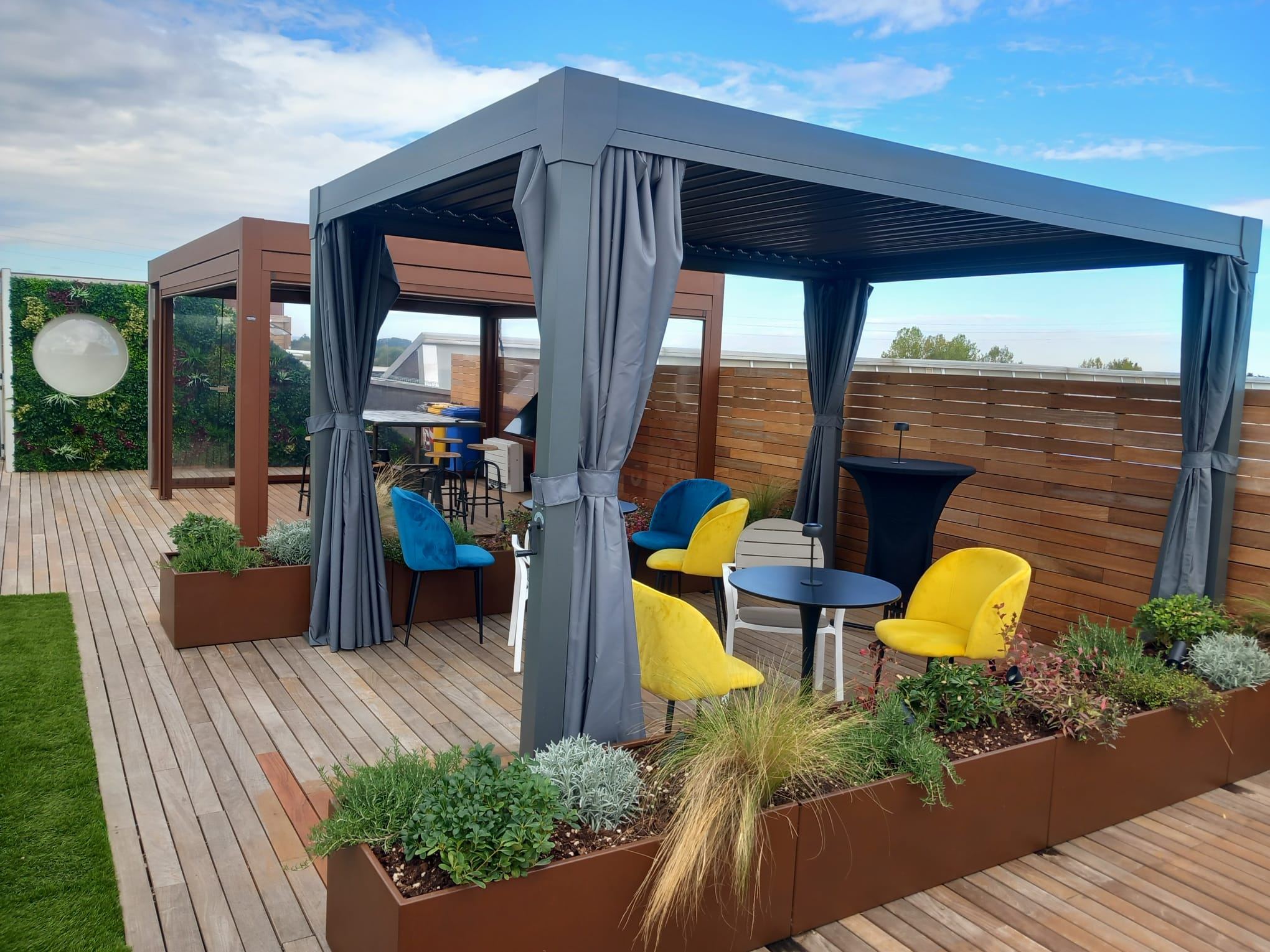 Rooftop aziendale con pergola, tende laterali, sedute gialle e fioriere su prato sintetico
